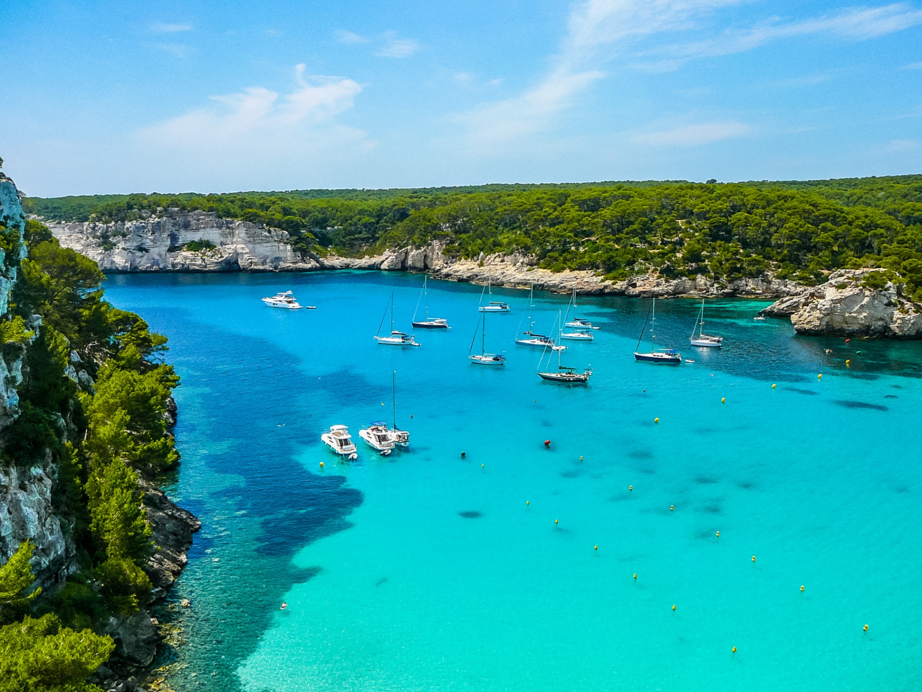 Fleet of sailing yachts and motor boats anchored between limestone cliffs in the turquoise waters of Cala Macarelleta, Menorca