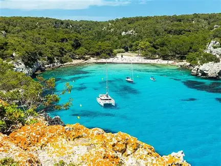 A catamaran and two yachts at anchor in Cala Algaiarens, a horseshoe cove backed by pine forest on Menorca's north-west coast
