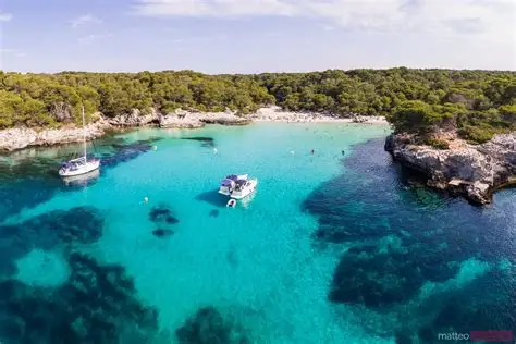 Two motor cruisers anchored in a small pine-fringed cove with clear turquoise water on Menorca's south coast